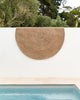 A round, brown rug by Baya NZ hangs over a wall by a swimming pool. 