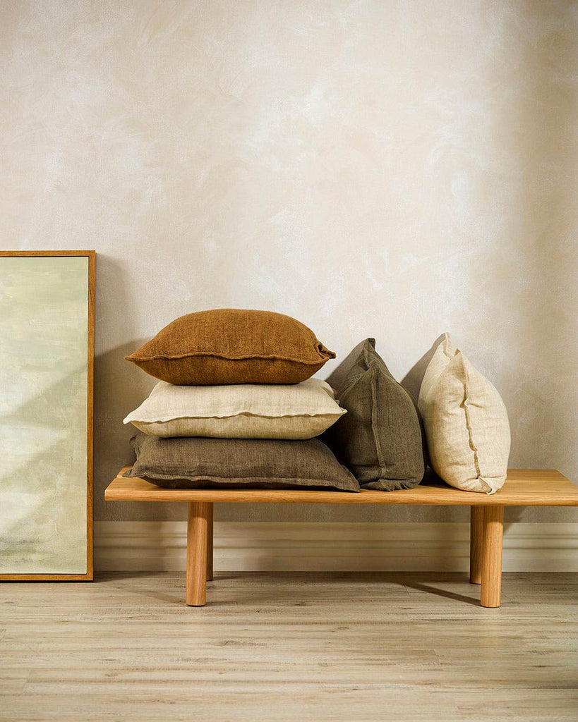 Baya Cassia Linen cushions stacked on a bench, including colours Clove Brown, Tobacco, and Almond