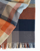 A folded 100% wool throw blanket with a colourful check plaid pattern in shades of orange, blue, and beige, and a twisted edge. 