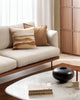 The corner of a couch and a coffee table, with a gorgeous feature cushion showcasing fine cotton printing with warm brown pigments. By Baya