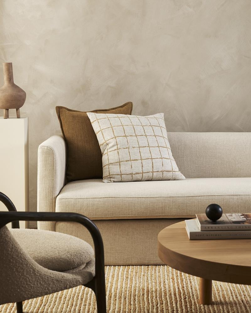 A natural linen-blend cushion with an ochre grid-look design in a hand-painted style, styled with a plain brown cushion in a stylish living room.