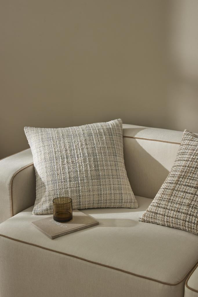 An on-trend woven cushion in white, alongside subtle beige and blue - the Tripoli Pearl by Weave, on a cream couch with brown binding.