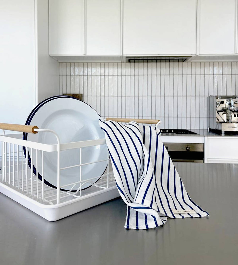 A striped tea towel from Father Rabbit, in the shade French Nautical Navy Stripe. It is draped over a dish rack, the blue and white stripes adding an element of whimsy to the kitchen.