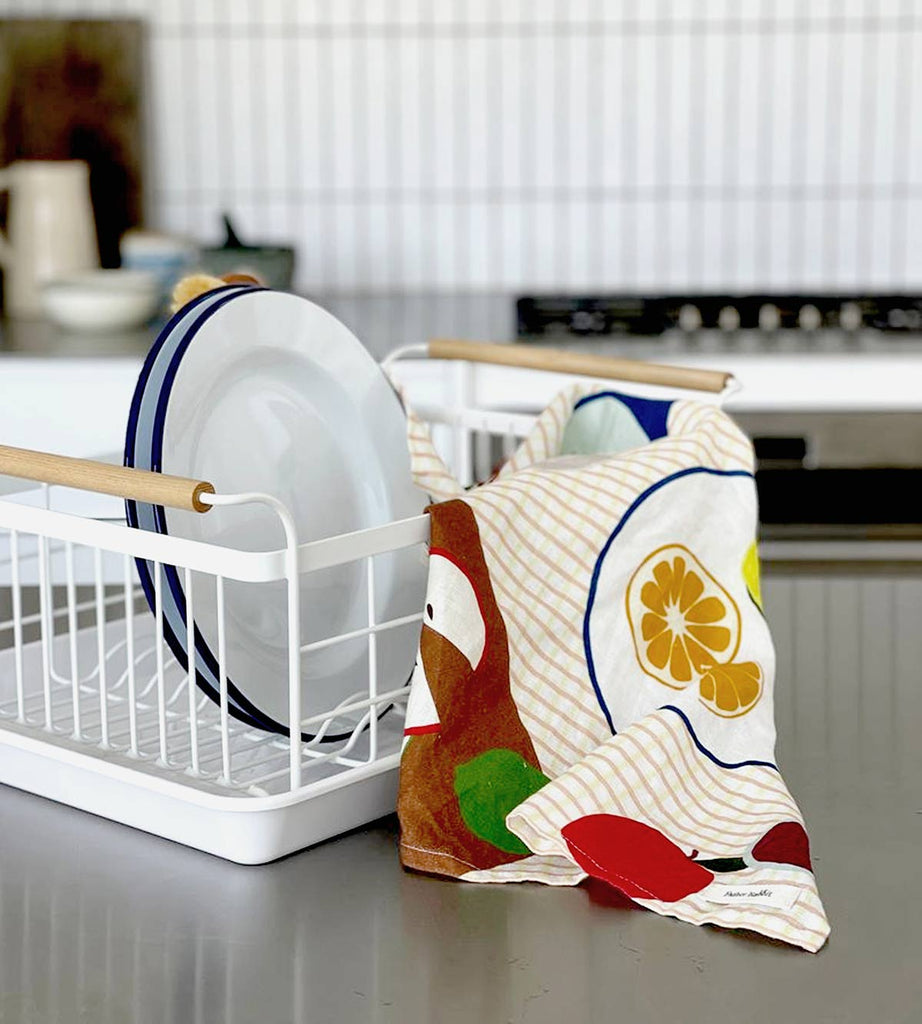 A Father Rabbit tea towel draped over a dish rack, featuring a playful 'Fruit on Plates' design. The towel shows vibrant fruit illustrations like apples and citrus, arranged on decorative plates, with a light checkered background.