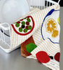 A Father Rabbit tea towel with the 'Fruit on Plates' pattern hangs over a dish rack, showcasing bright fruit motifs such as apples and citrus slices on plate illustrations. The towel has a subtle checkered background, complementing the white plates with blue rims in the drying rack, creating a fresh, lively kitchen atmosphere.