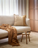 A textural cotton cushion with stripe detailing in warm brown tones, on a cream couch with a caramel brown throw blanket.