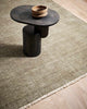 A coffee table on top of a lightly distressed olive green floor rug, with white detailing including a subtle fringe, in 100% NZ wool. 