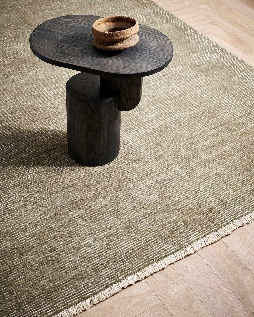 A coffee table on top of a lightly distressed olive green floor rug, with white detailing including a subtle fringe, in 100% NZ wool. 