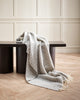 A soft 100% wool throw blanket draped over a designer chair, with silver striping and cream twisted edging. 