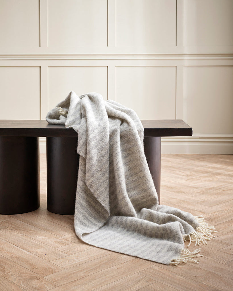 A soft 100% wool throw blanket draped over a designer chair, with silver striping and cream twisted edging. 
