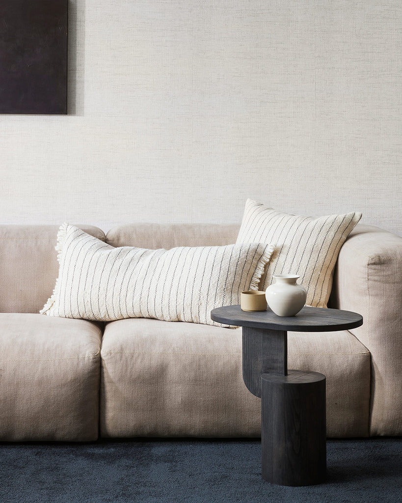 A modern living room with two cushions on the couch, both with the same pattern. One is a long lumbar and one is square, and they are made of the same artisanal handwoven cotton with a linear pattern in navy blue against soft cream. 
