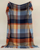 A person holding a large, 100% wool throw blanket up. The blanket has a striking vintage style check pattern in shades of brown, red, and blues. 