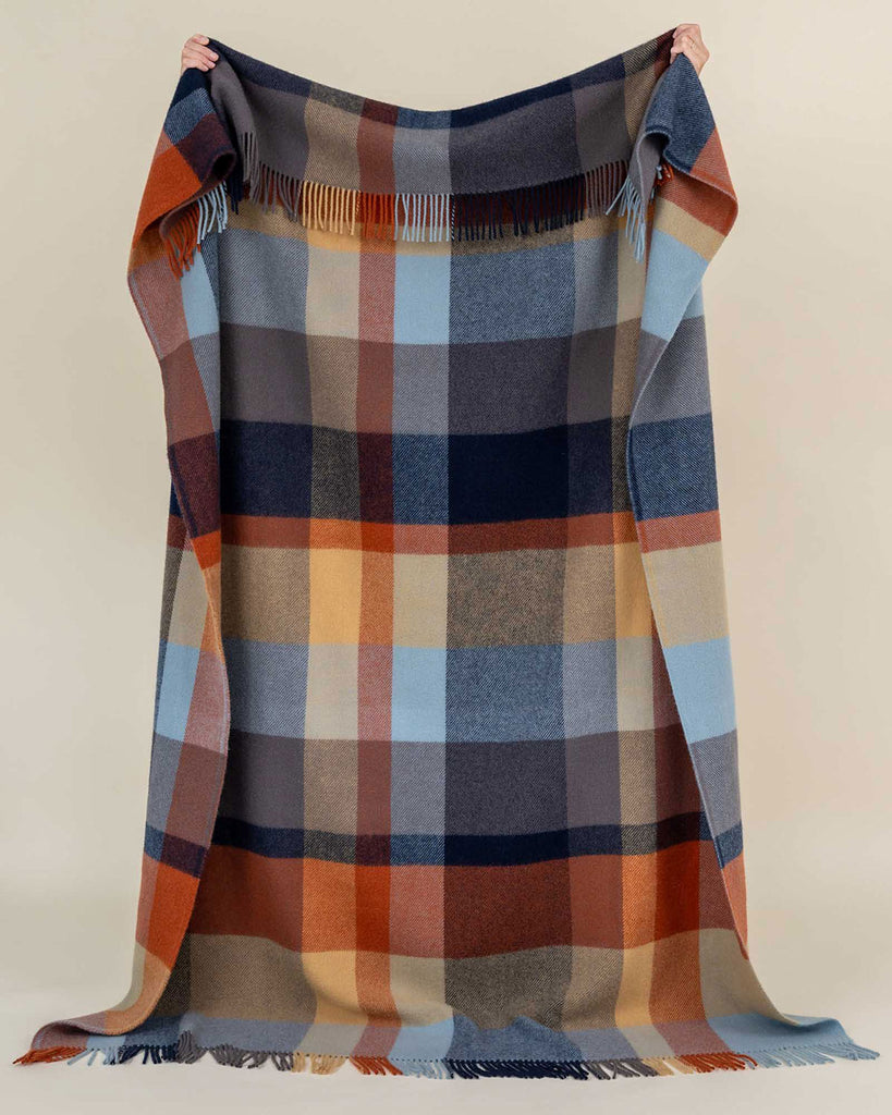 A person holding a large, 100% wool throw blanket up. The blanket has a striking vintage style check pattern in shades of brown, red, and blues. 