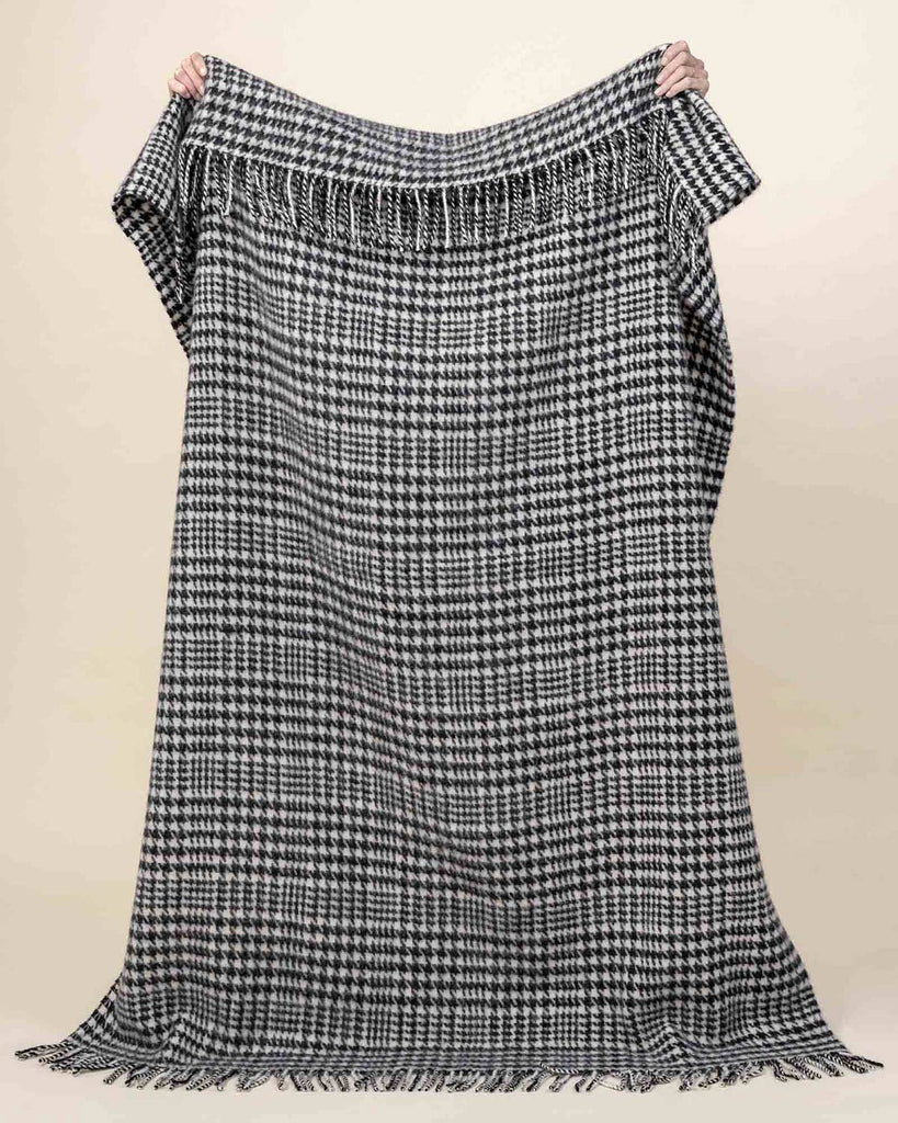 A full image of the Helmsdale, a black and white houndstooth patterned throw blanket with fringed edging in 100% wool, held up to show the generous sizing. 