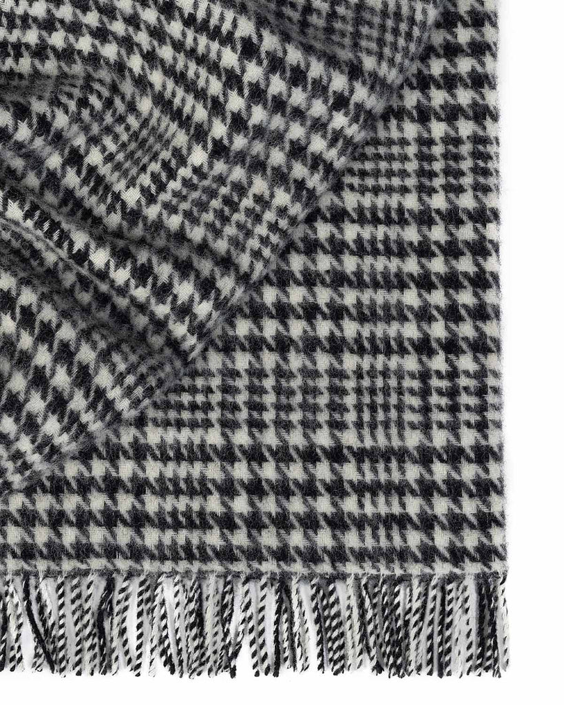 A folded throw blanket in 100% wool with a sophisticated black and white houndstooth check pattern and twisted fringe edging.
