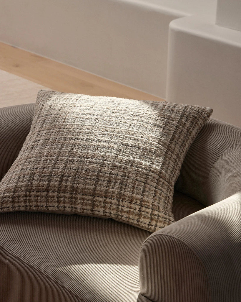 A woven textural cushion - the Tripoli by Weave Home, in warm ivory and brown tones, on a chair in a light, sunny room