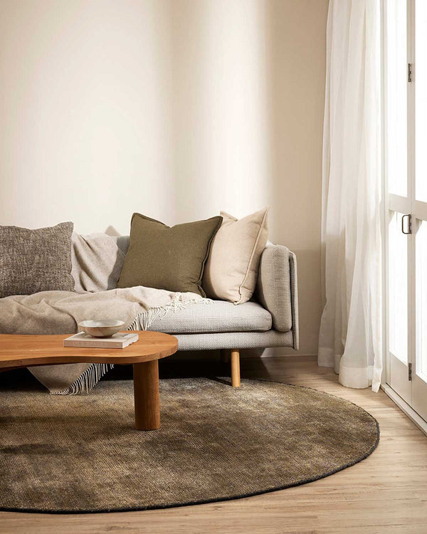 A round, earthy grey rug with a couch and cushions by Baya NZ situated in a stylish living room. 