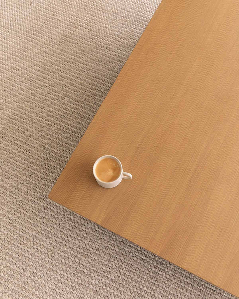 A top view of a simple table with a coffee on it. Underneath the table is a woven brown rug by Baya NZ