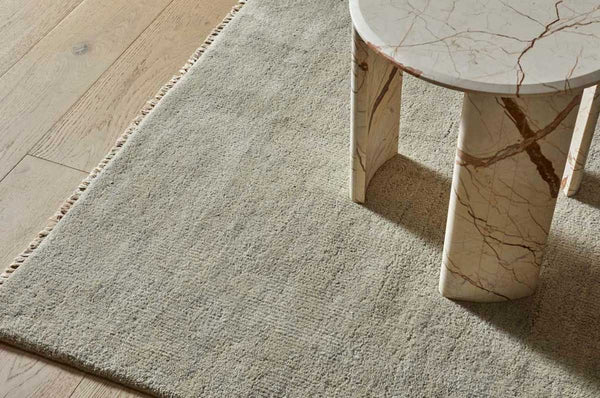 A soft, handloomed rug made of 100% wool, in a soft beige ivory tone by Soren Liv. Situated under a table.