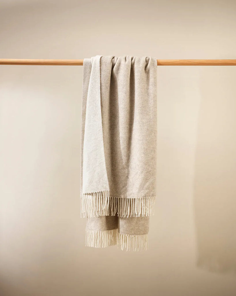 Merino-blend throw blanket with reversible cream and oatmeal tones, hanging with tasselled fringe