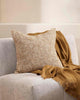 A textural woven cushion in shades of muted mustard yellow and ercu, styled on a cream armchair with a mustard brown throw blanket by Baya