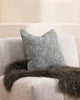 A textural cushion in shades of sage green and ercu, with a prominent weave, by Baya, styled with a fashionable faux fur throw on a designer armchair