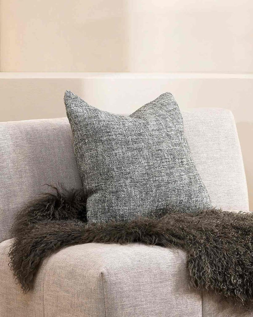A textural cushion in shades of sage green and ercu, with a prominent weave, by Baya, styled with a fashionable faux fur throw on a designer armchair