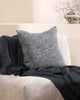 A textural woven cushion in shades of slate, blue-black and ercu, styled on a modern armchair with a complimentary blue throw blanket by Baya