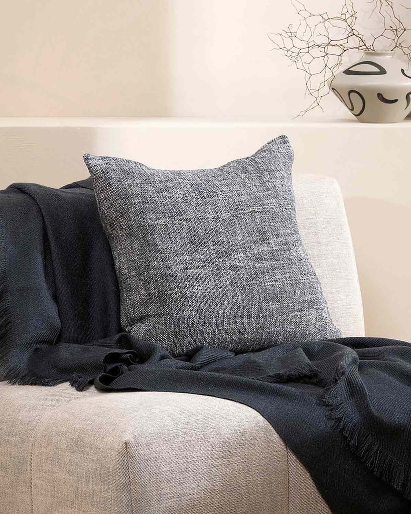A textural woven cushion in shades of slate, blue-black and ercu, styled on a modern armchair with a complimentary blue throw blanket by Baya