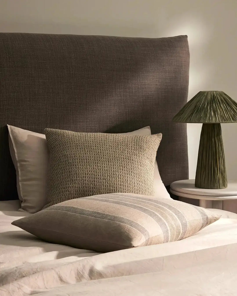 A neutral, textural cushion by Weave Home in a natural linen colour, seen on a bed paired with a complemetary plain and a stripe