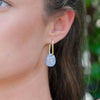 A woman wearing a dainty pair of gold square hoops with an iridescent blue acrylic charm with a sweet star etched into it. 