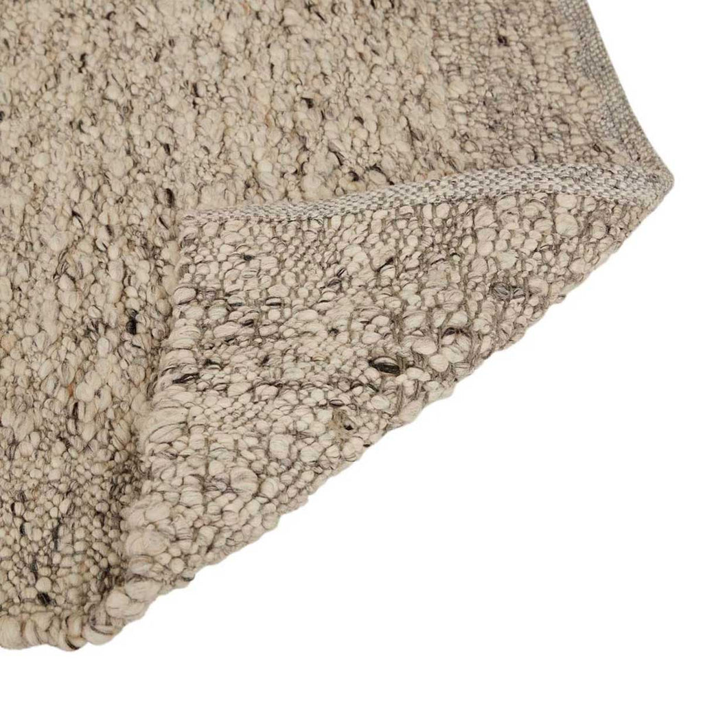 Close up of the weave of a textural wool blend boucle style rug by Globe West and Soren Liv NZ in shades of light grey and brown. The edge of the rug is flipped, revealing the underside. 