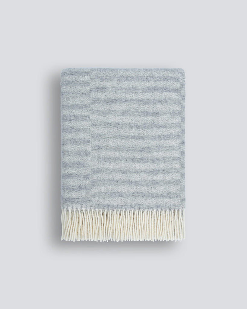 An off-set striped patterned throw blanket in shades of cool silver and pale blue, with a twisted fringe edge in 100% NZ wool by Baya