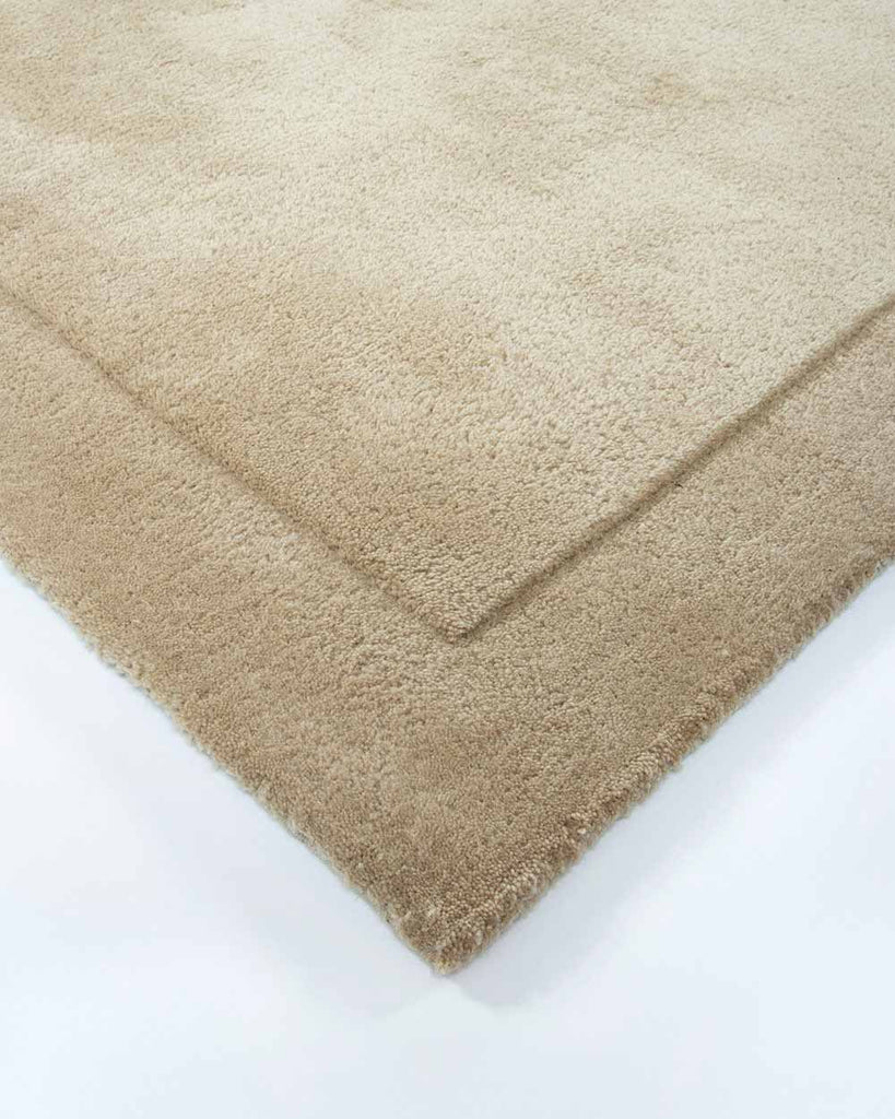 A corner view of the luxurious Norfolk rug in the shade Sand, a warm beige. The cut-pile border lends a sense of minimalism to this sumptous, 100% wool floor rug. 