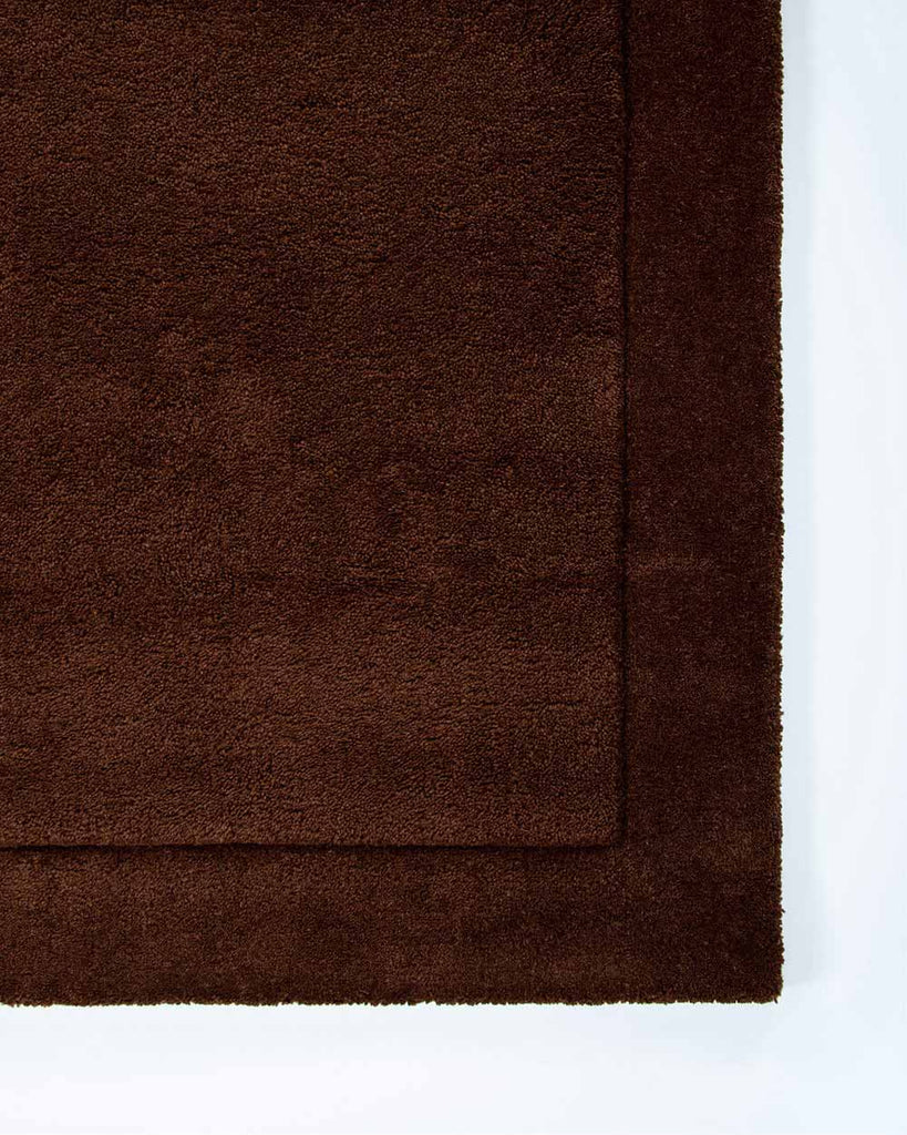 The corner of the Norfolk floor rug in the shade Chocolate, viewed from above. The border of the rug is cut-pile in a minimalist and refined design. 