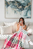 A woman sitting on a couch in a stylish living room enjoying the morning sun with a cup of coffee. Across her knees is a pink checked throw blanket by Ruanui Station, NZ. 