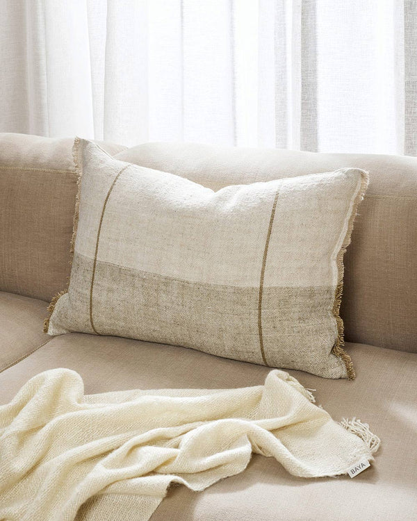 A lumbar cushion in textural linen with tones of warm beige, sandy brown, and cream, with fringed edges on a beige toned couch. 