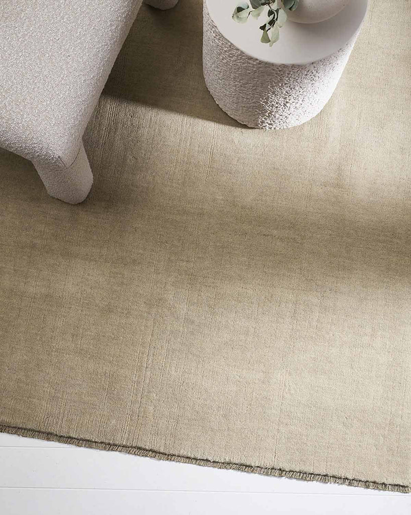 A soft beige floor rug seen from above with a designer couch and coffee table, fringed edges, and 100% wool. 