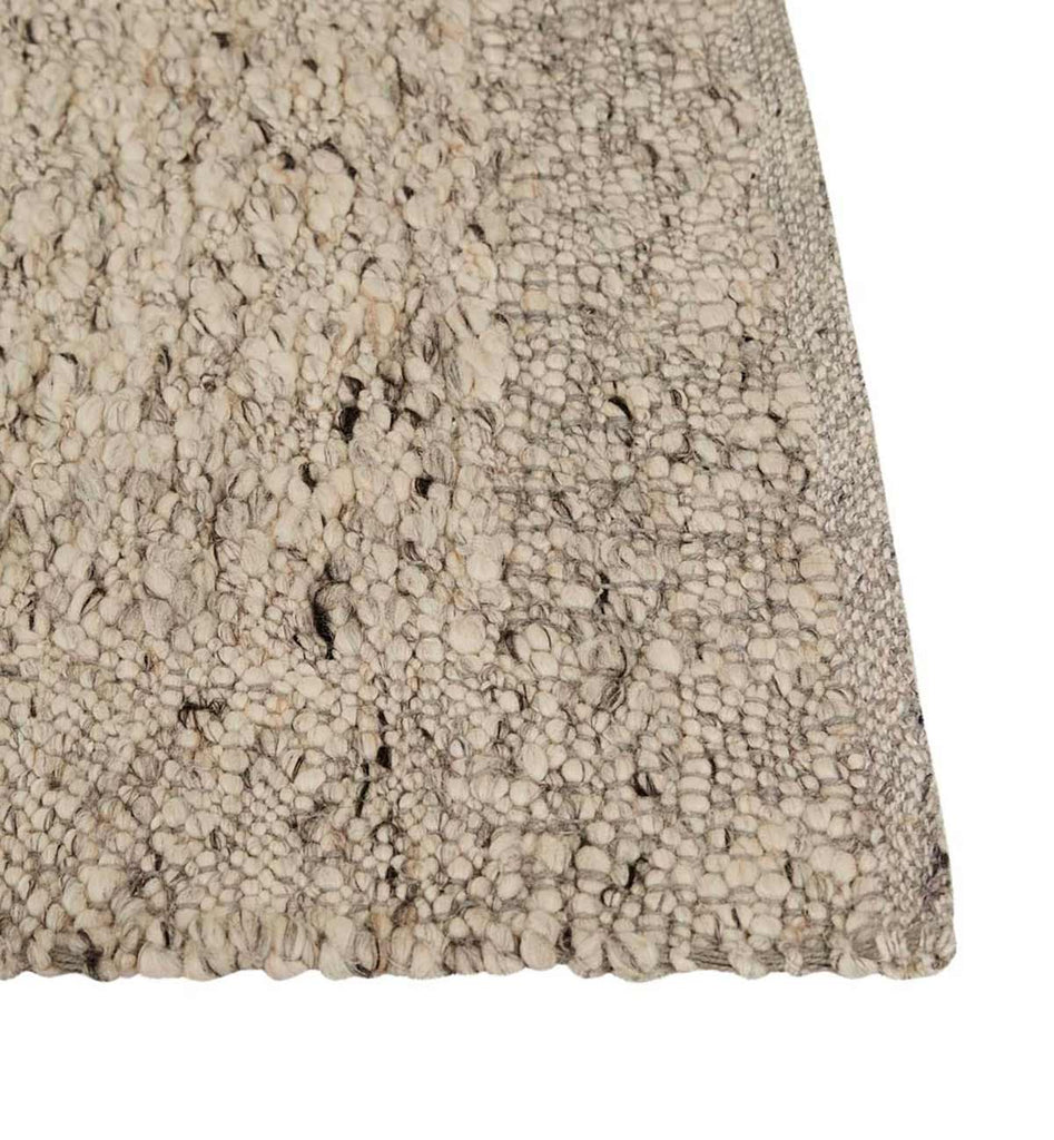 Close up of the corner of a textural wool blend boucle style rug by Globe West and Soren Liv NZ in shades of light grey and brown. The edge of the rug is indented, showing interest to the rug 